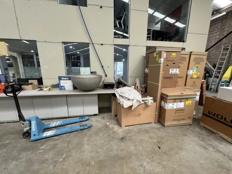 Stacked cartons, workshop furniture and a blue pallet jack beside the internal office area.