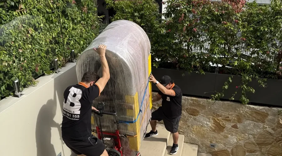 Two JB Removals specialists guiding a fully protected 250kg ice bath vertically up an outdoor staircase in Vaucluse